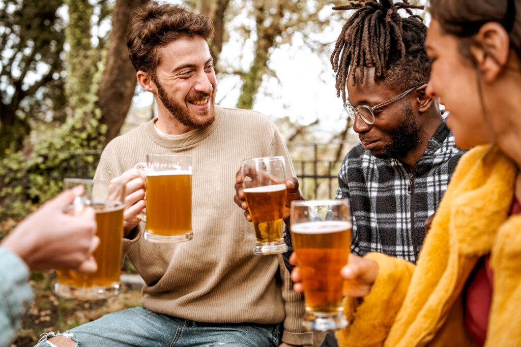 Multiethnic group of young friends having a party in the forest pub having fun talking and drinking beers - alcohol drink, diversity, wellbeing and people lifestyle concept