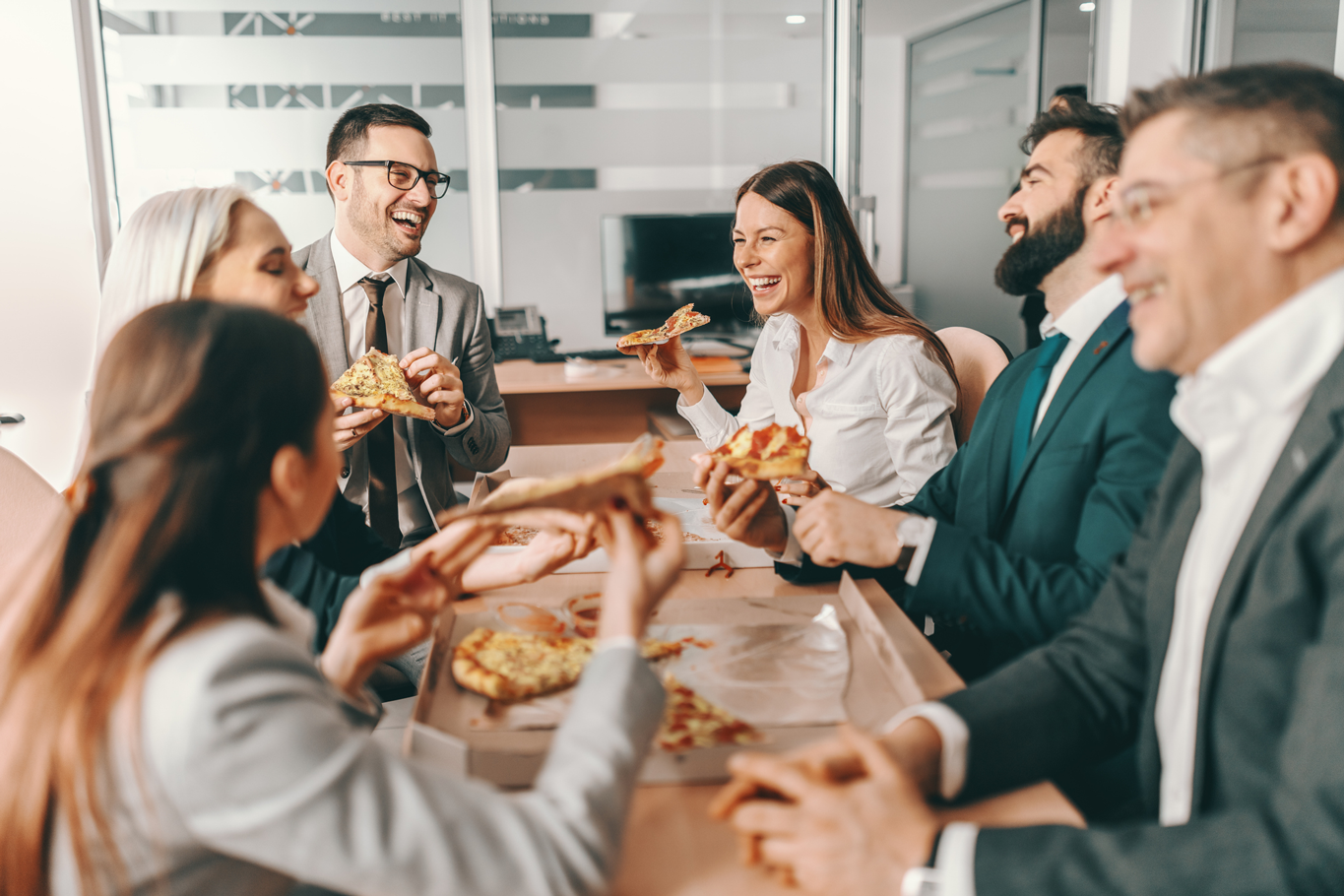 Small group of happy colleagues in formal wear chatting and eating pizza together for lunch.