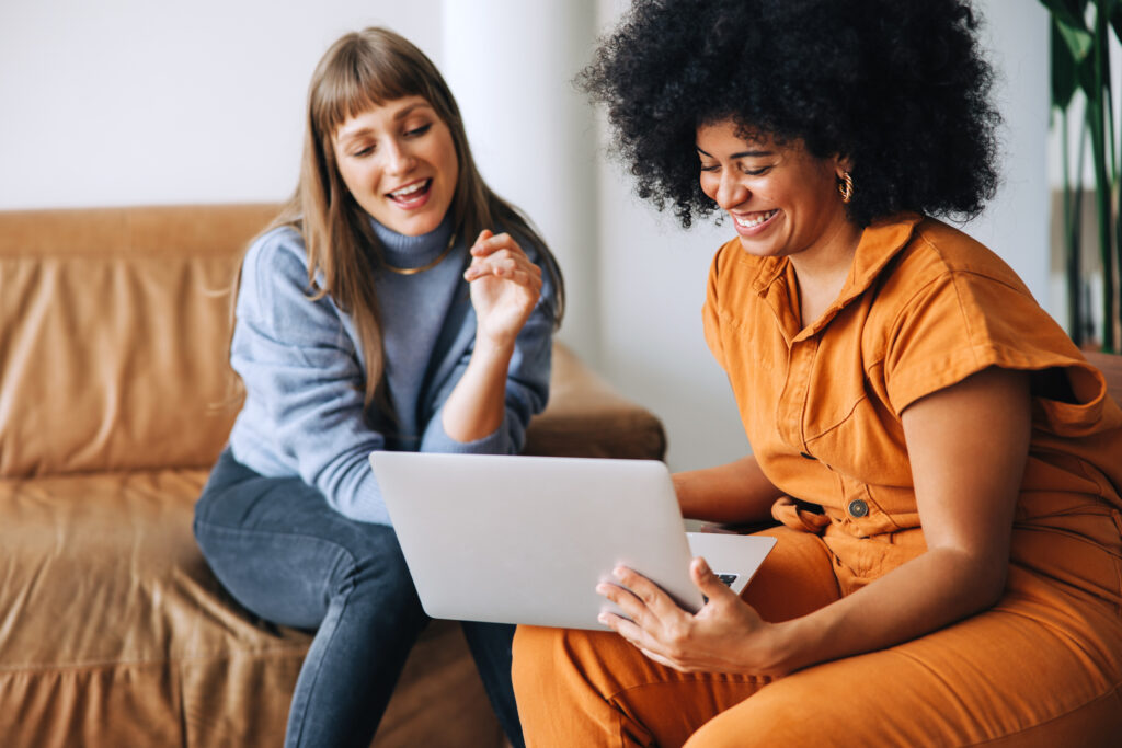 Cheerful young businesswomen smiling happily while using a laptop together. Two female entrepreneurs working as a team in a modern workplace.