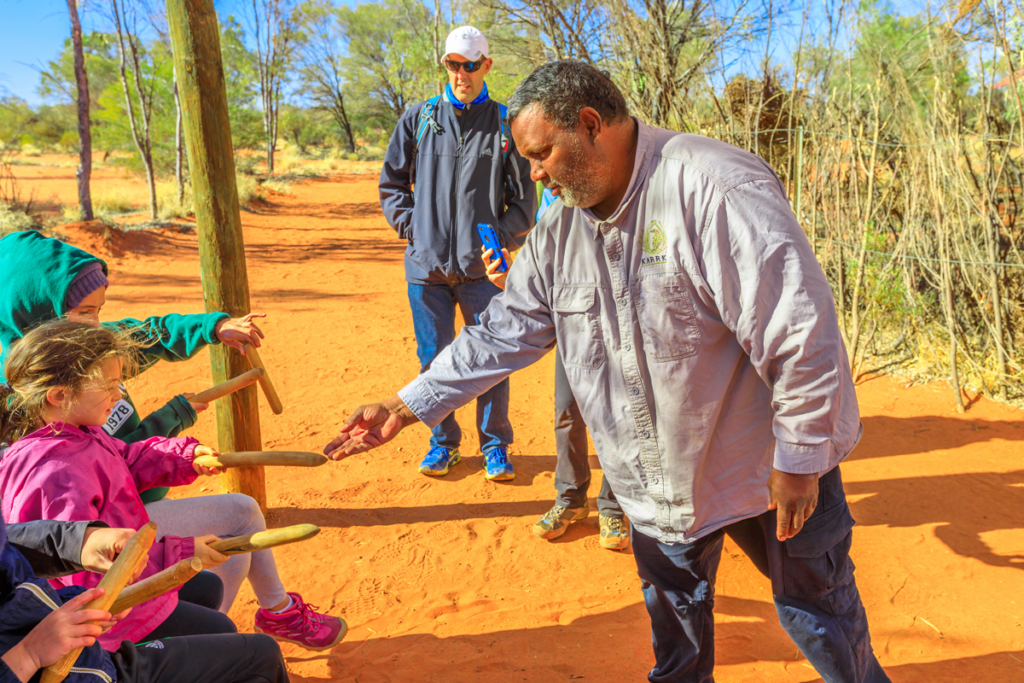 A man demonstrating Aboriginal instruments to children in rural bushland of Australia.