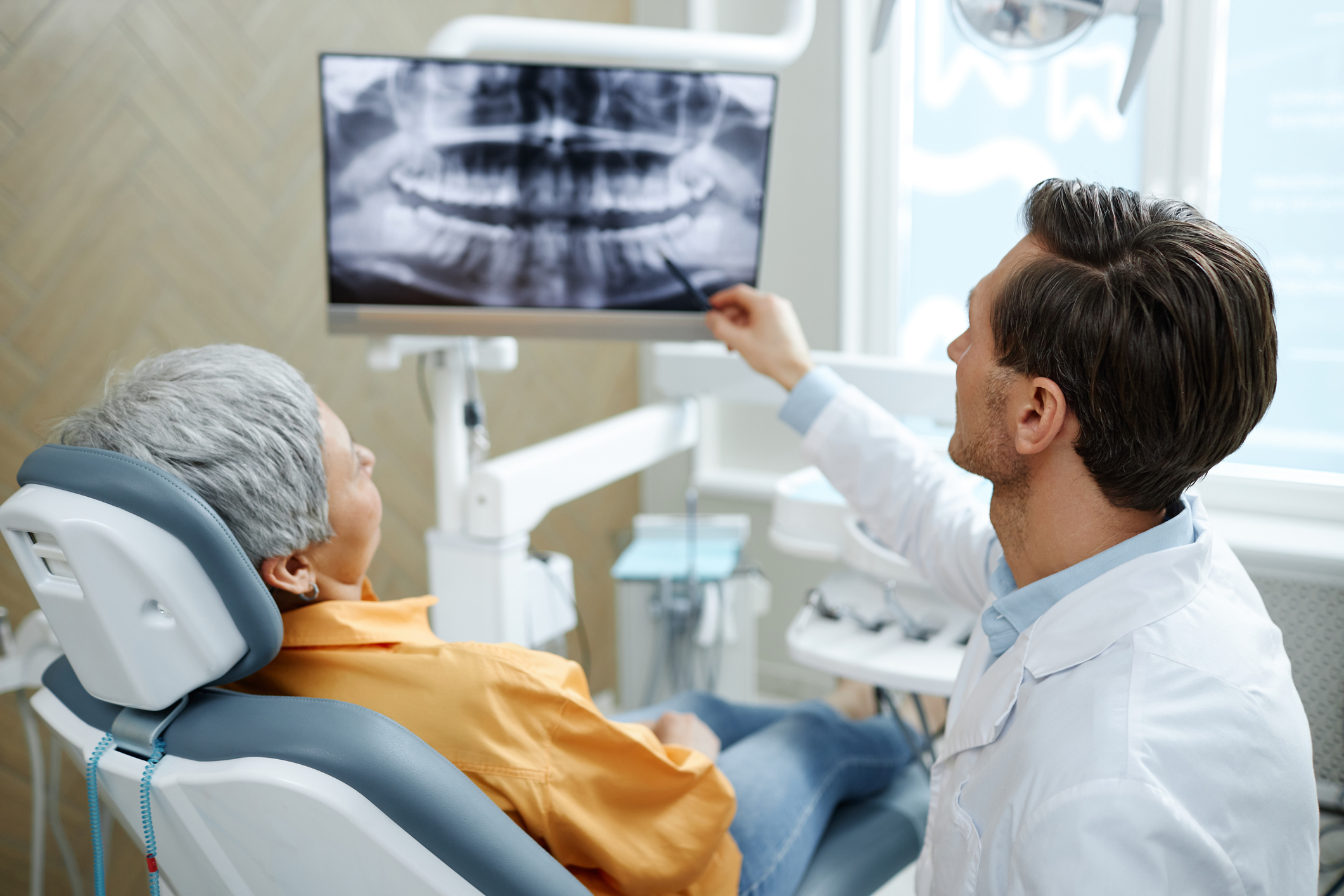 Back view of male dentist pointing at tooth X-ray image on screen during consultation in modern dental clinic