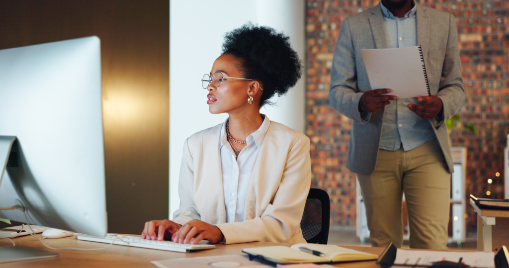 A woman focused on her computer screen while a man stands behind her, looking over her shoulder.