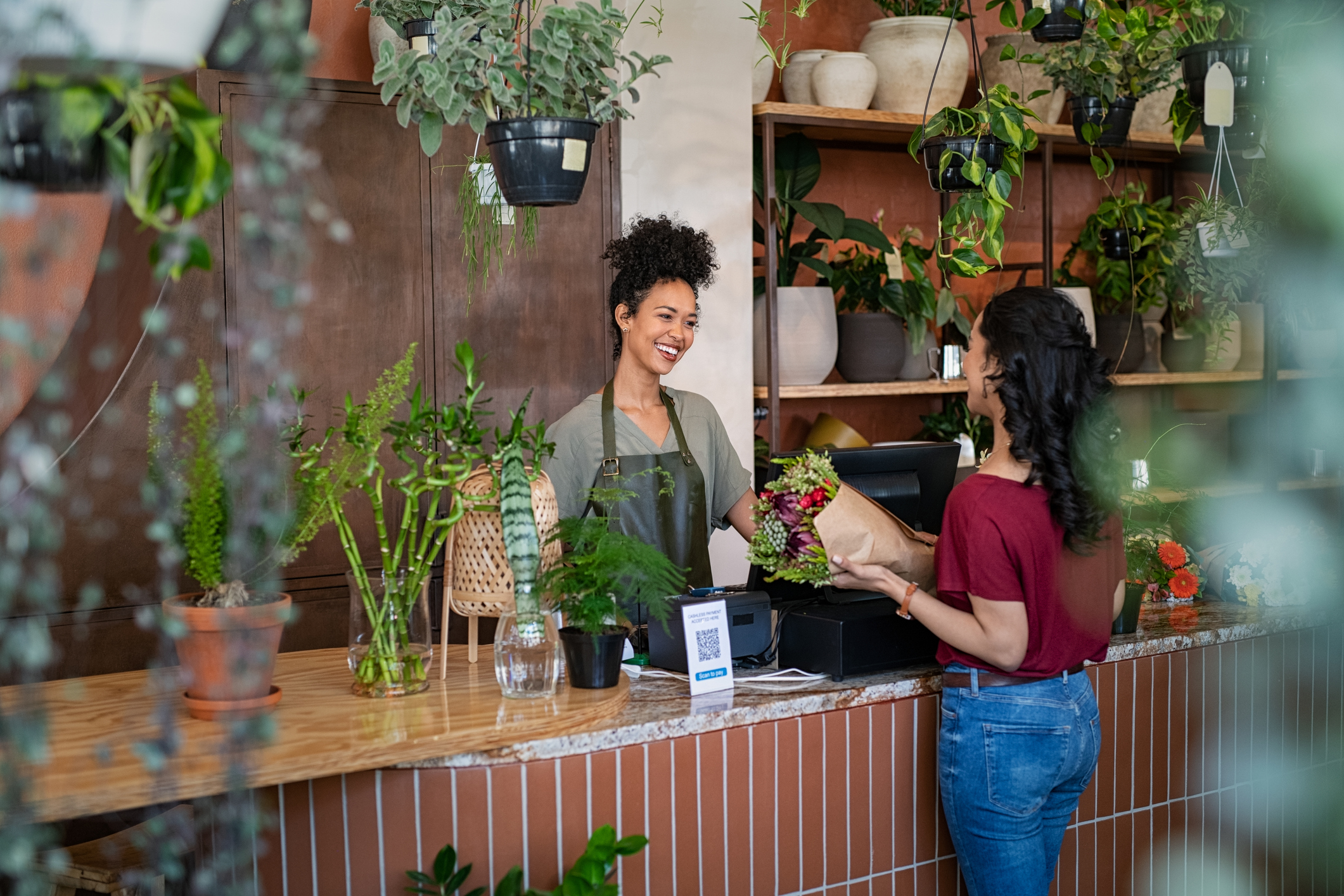 A woman standing at a counter, smiling, next to a vibrant green plant in a pot.