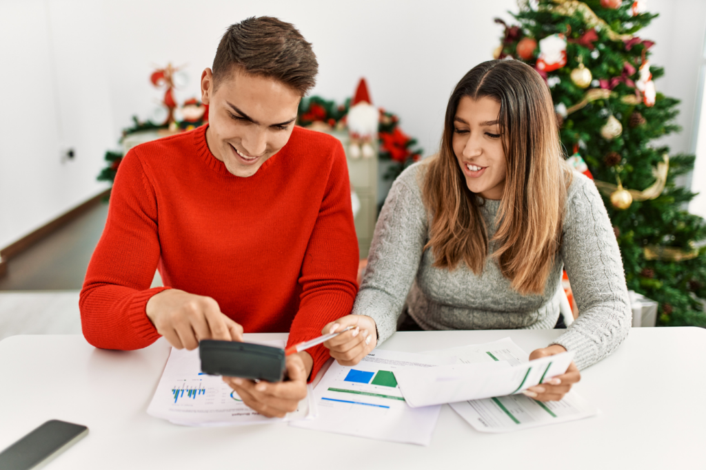 A young couple sitting at a table with Christmas decorations, both looking at their phone with smiles.