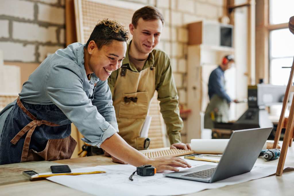 Two individuals collaborating on a laptop amidst woodworking tools and materials in a workshop.