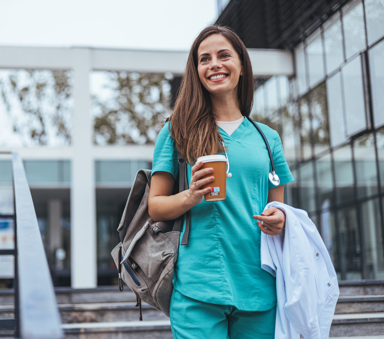 The young adult female nurse medical doctor drinks her coffee as she arrives to work at the hospital. A young adult female nurse arrives for a shift in a hospital. She is wearing medical scrubs