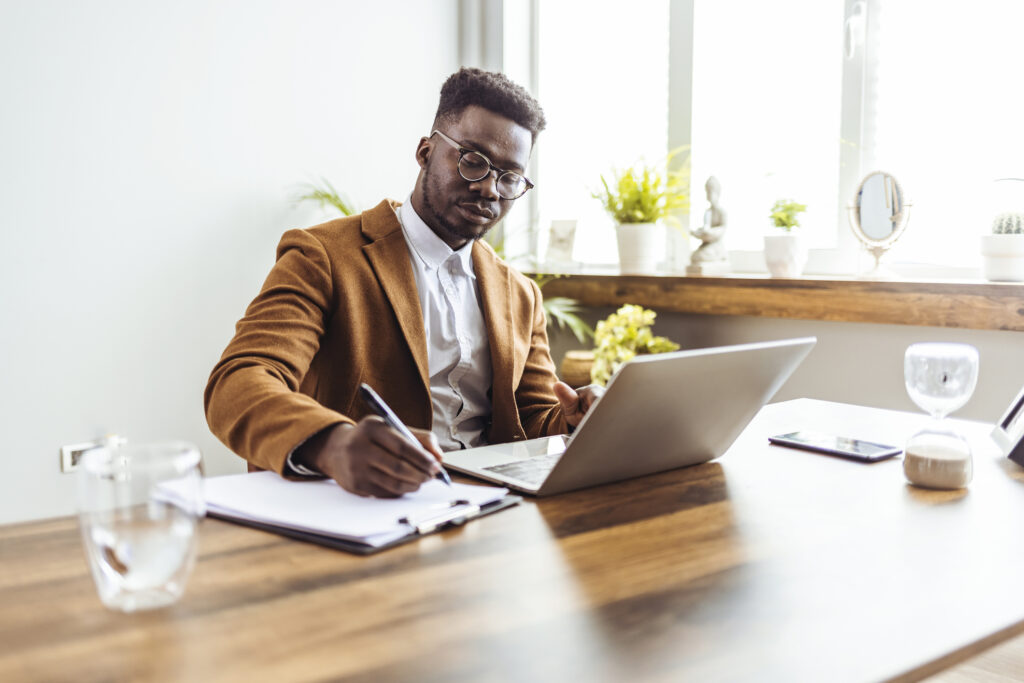 Concentrated African man in glasses sit at desk work on computer make notes. Focused serious male worker or freelancer busy at home office watch webinar on laptop, handwrite on paper.