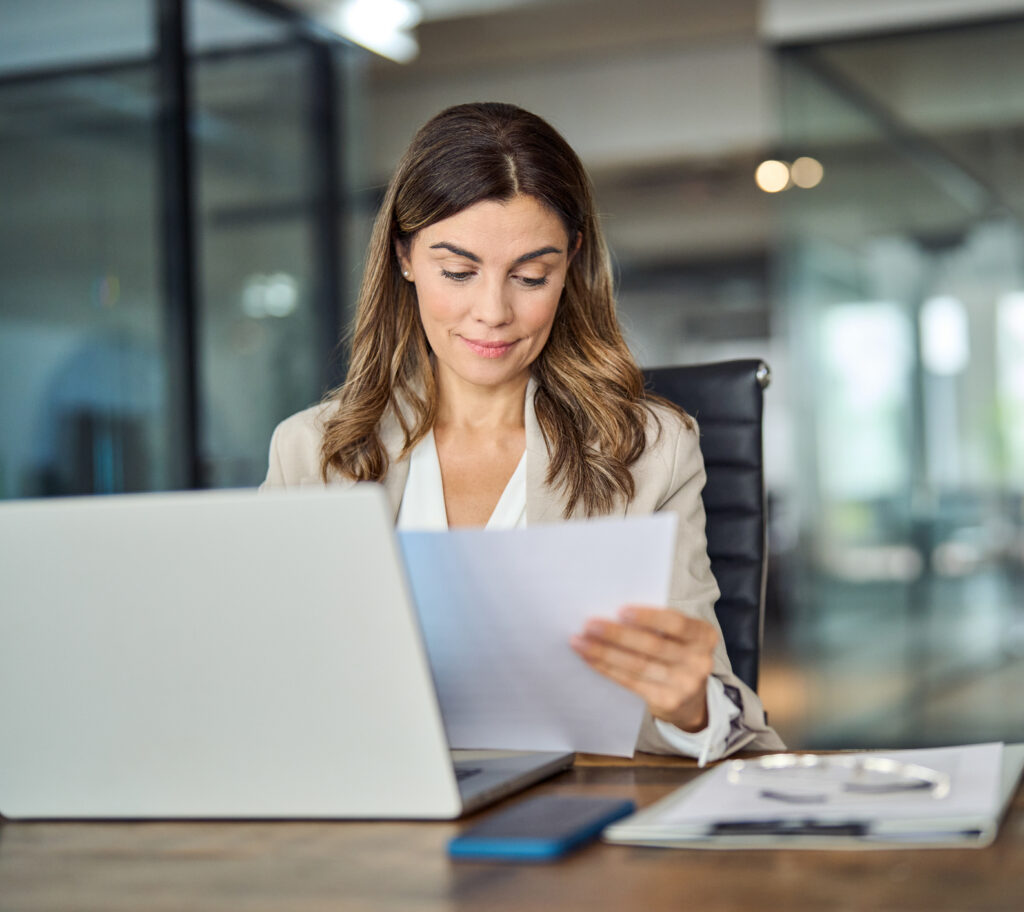 Busy mid aged business woman working in office with laptop reading document. Mature professional female manager lawyer attorney holding paper finance report sitting at desk in office. Lawyer office business