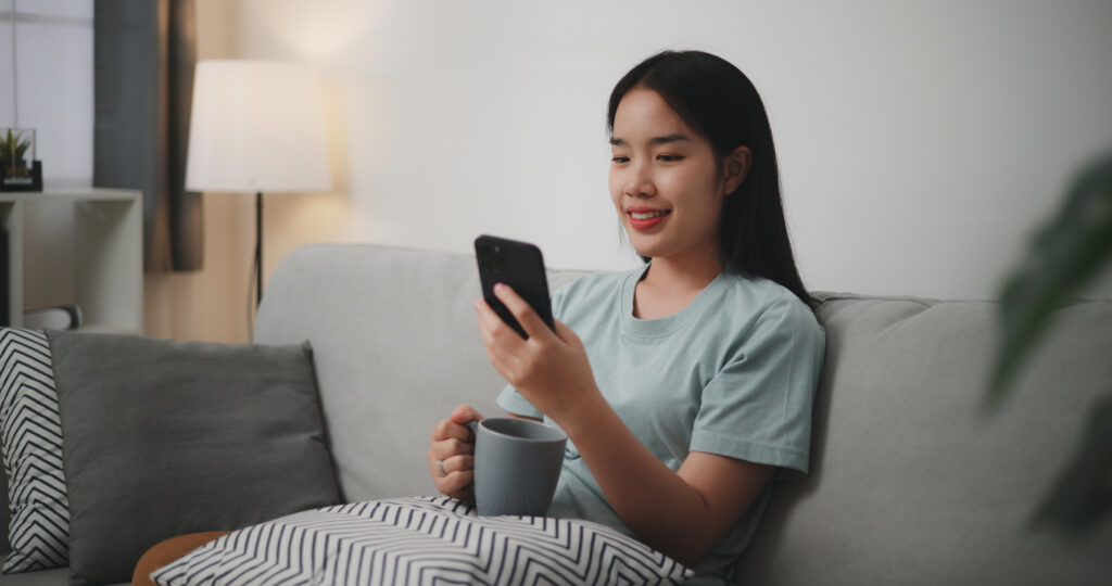 a woman checking her phone on the couch with a cup of tea in hand, smiling at phone